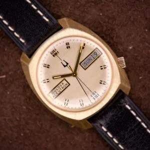 A Bulova Accutron Turning Fork Cal. 2182 Day-Date Cushion Case wristwatch with a gold finish, black leather strap, and iconic tuning fork movement, featuring a dial that shows both day and date.