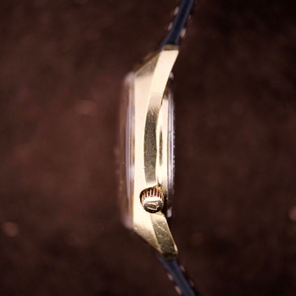 Close-up side view of a gold Bulova Accutron Turning Fork Cal. 2182 Day-Date Cushion Case wristwatch with a textured crown, set against a brown background.