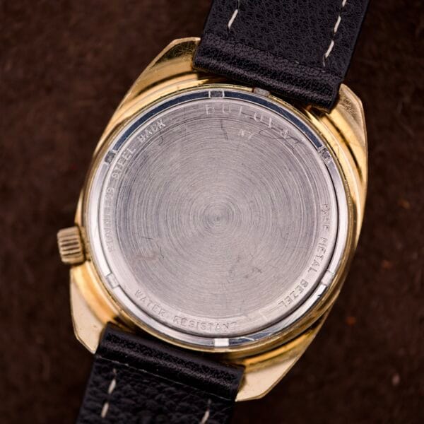 The back of a vintage Bulova Accutron Turning Fork Cal. 2182 Day-Date Cushion Case wristwatch features a black leather strap and engraved text on the metal case.