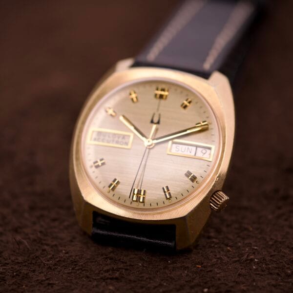 Bulova Accutron Turning Fork Cal. 2182 Day-Date Cushion Case wristwatch with black leather strap, displayed on a brown textured surface.