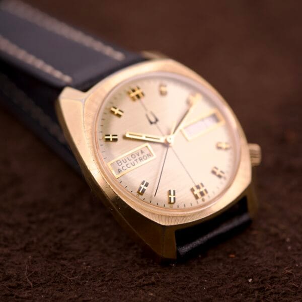 A Bulova Accutron Turning Fork Cal. 2182 Day-Date Cushion Case with a black leather strap rests on a brown textured surface.