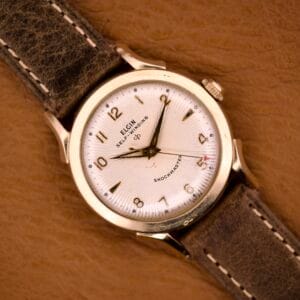 Close-up of an Elgin Automatic Shock-Master Guilloché Dial Cal. 643 wristwatch with a brown leather strap, shown on a tan background.