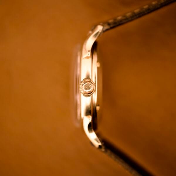 Close-up side view of the Elgin Automatic Shock-Master Guilloché Dial Cal. 643 gold watch crown against a blurred brown background.
