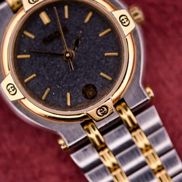 Close-up of the Gucci 9000 Ladies Watch with a black slate dial and two-tone quartz band, shown against a striking red background.