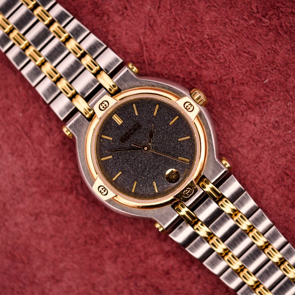 A Gucci 9000 Ladies Watch with a black slate dial and two-tone quartz design, displayed on a textured red surface.