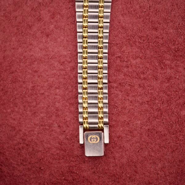 Close-up of the Gucci 9000 Ladies Watch with a black slate dial and two-tone quartz band, showcased against a textured reddish background.