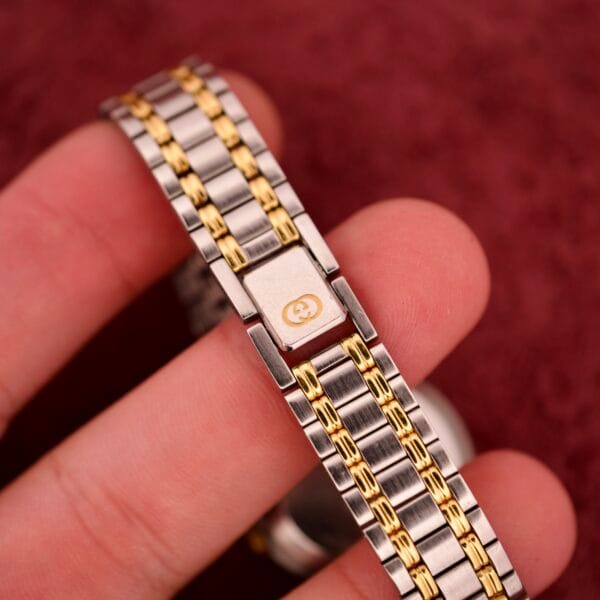 Close-up of a hand holding a Gucci 9000 Ladies Watch Black Slate Dial Two-Tone Quartz, showing the logo on the clasp, against a red background.