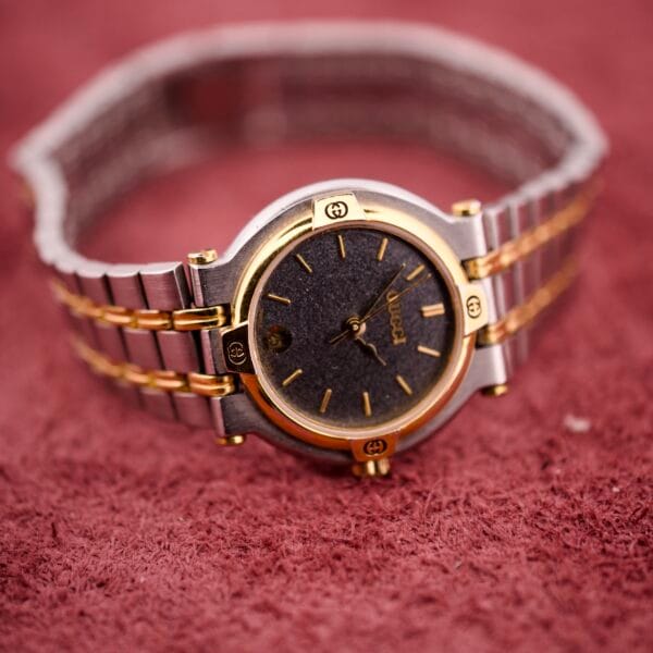 The Gucci 9000 Ladies Watch features a black slate dial and two-tone quartz design, showcased elegantly on a textured red surface.