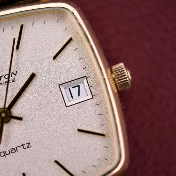 Close-up of a Hamilton Masterpiece Ref. 9116 10K Gold Filled TV Case Quartz watch with a white face displaying the date "17," set against a maroon background. The 10K gold filled case lends sophistication to this elegant timepiece.