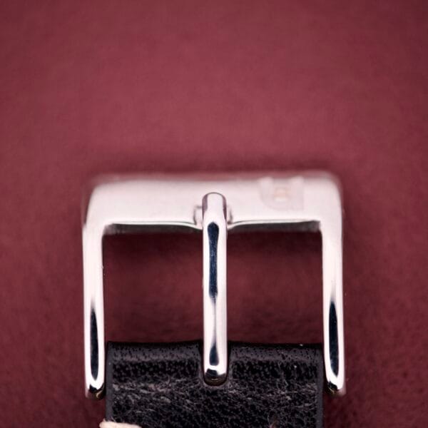 A close-up of a silver buckle on black leather against maroon, evoking the refined elegance of the Hamilton Masterpiece Ref. 9116 10K Gold Filled TV Case Quartz.