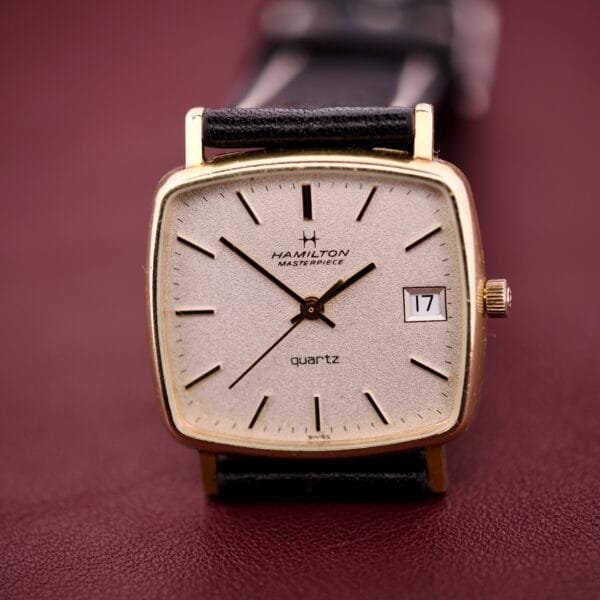 The Hamilton Masterpiece Ref. 9116 features a 10K gold-filled TV case, quartz movement, date display on a maroon dial, and a black strap.