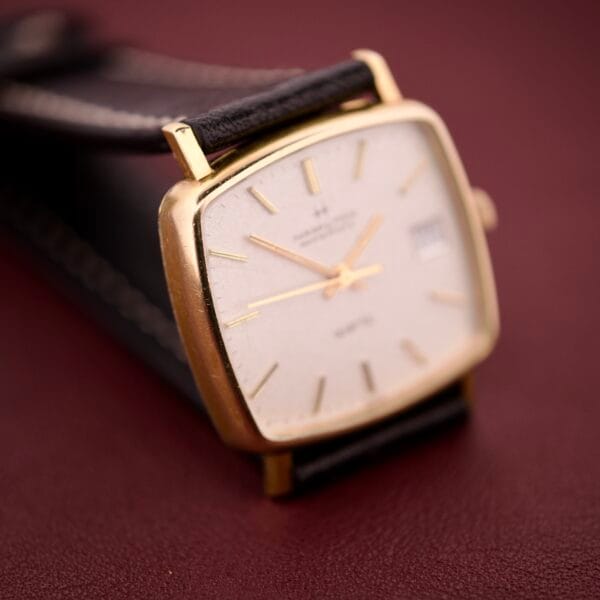 A Hamilton Masterpiece Ref. 9116 10K Gold Filled TV Case Quartz wristwatch with a leather strap rests on a dark red surface.