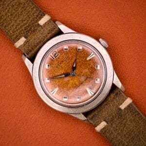 Benrus Shock-Absorber Tropic Dial wristwatch with silver indices, brown face, and green leather strap, displayed on an orange background.