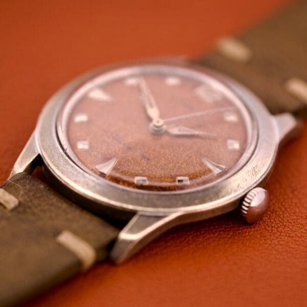 Close-up of a vintage Benrus Shock-Absorber Tropic Dial Silver Indicies wristwatch with a brown leather strap, displayed on an orange surface.