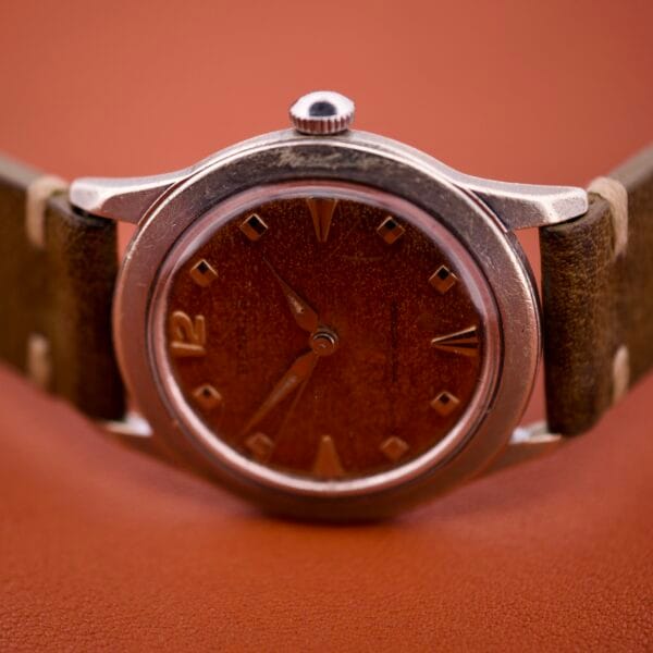 The Benrus Shock-Absorber Tropic Dial wristwatch features silver indices, a brown face, and a leather strap, displayed against a reddish-brown background.