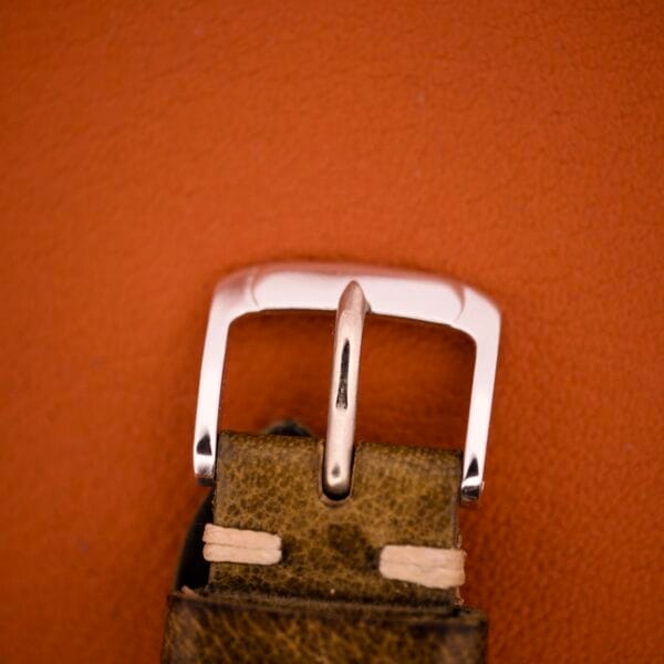 A close-up of a worn brown leather strap with a silver buckle on an orange background complements the vintage Benrus Shock-Absorber Tropic Dial Silver Indicies and would pair well with its classic design.