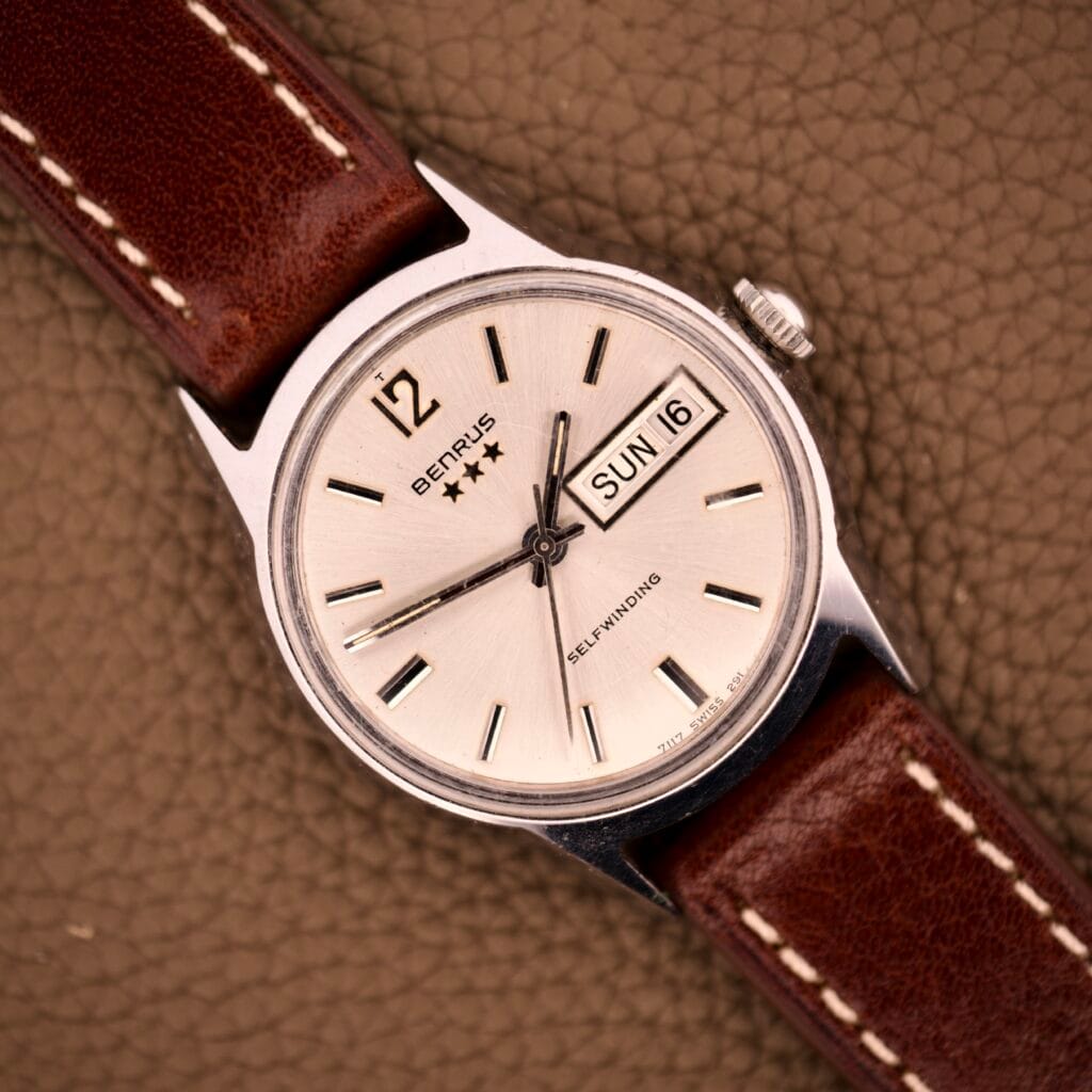 The Benrus Three Star Automatic Day-Date Sunburst Dial #7001 Series wristwatch features a silver case, brown leather strap, and displays day and date on its textured brown sunburst dial.