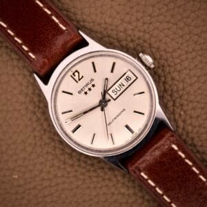 The Benrus Three Star Automatic Day-Date Sunburst Dial #7001 Series wristwatch features a silver case, brown leather strap, and displays day and date on its textured brown sunburst dial.