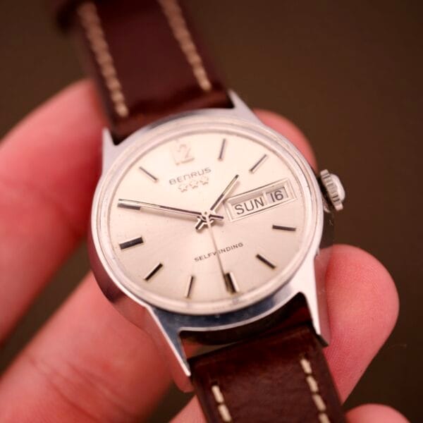 A hand holds the Benrus Three Star Automatic Day-Date Sunburst Dial #7001 Series wristwatch with a brown leather strap, proudly displaying Sunday 16.