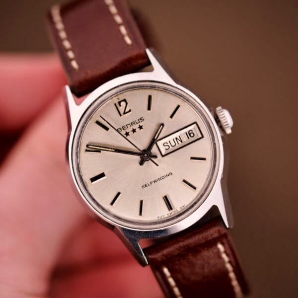 A hand holding the Benrus Three Star Automatic Day-Date Sunburst Dial #7001 Series wristwatch with a brown leather strap.