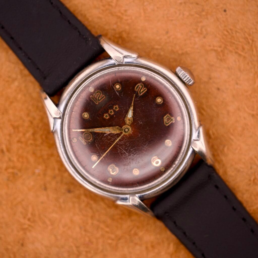 The Benrus Three Star Automatic features a browned tropic dial, stainless steel case, gold markers, and a black leather strap, combining vintage style with timeless appeal. Displayed on a tan surface.