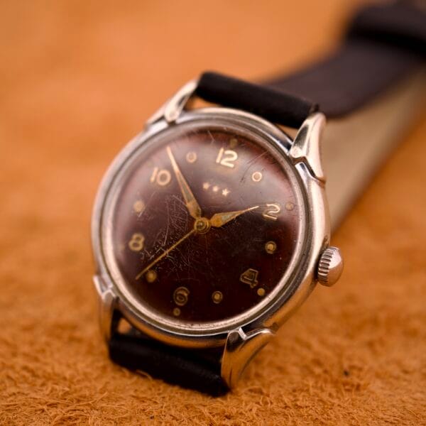 A Benrus Three Star Automatic wristwatch with a browned tropic dial and stainless steel case rests on a textured brown surface.