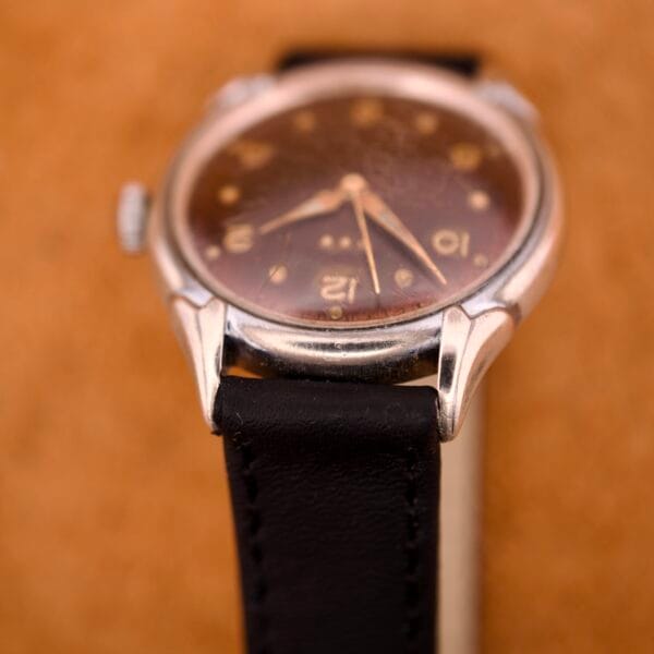 Close-up of a Benrus Three Star Automatic Browned Tropic Dial Stainless Steel wristwatch with a black leather strap, set against a brown background.