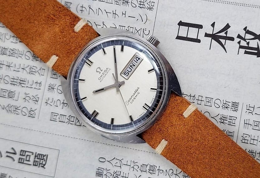 A wristwatch with a brown strap, likely an Omega Seamaster Cosmic, rests on a Japanese newspaper, displaying the time and day SUN 14.