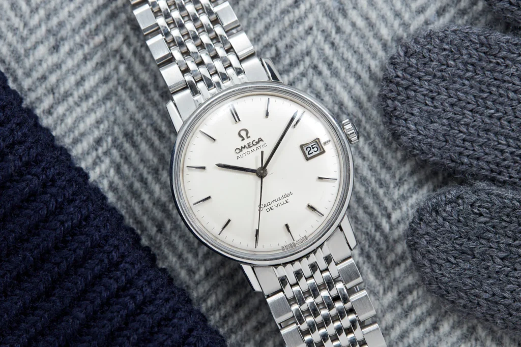 A silver Omega Seamaster Deville watch with a white face rests on gray and white striped fabric, beside elegant gray gloves.