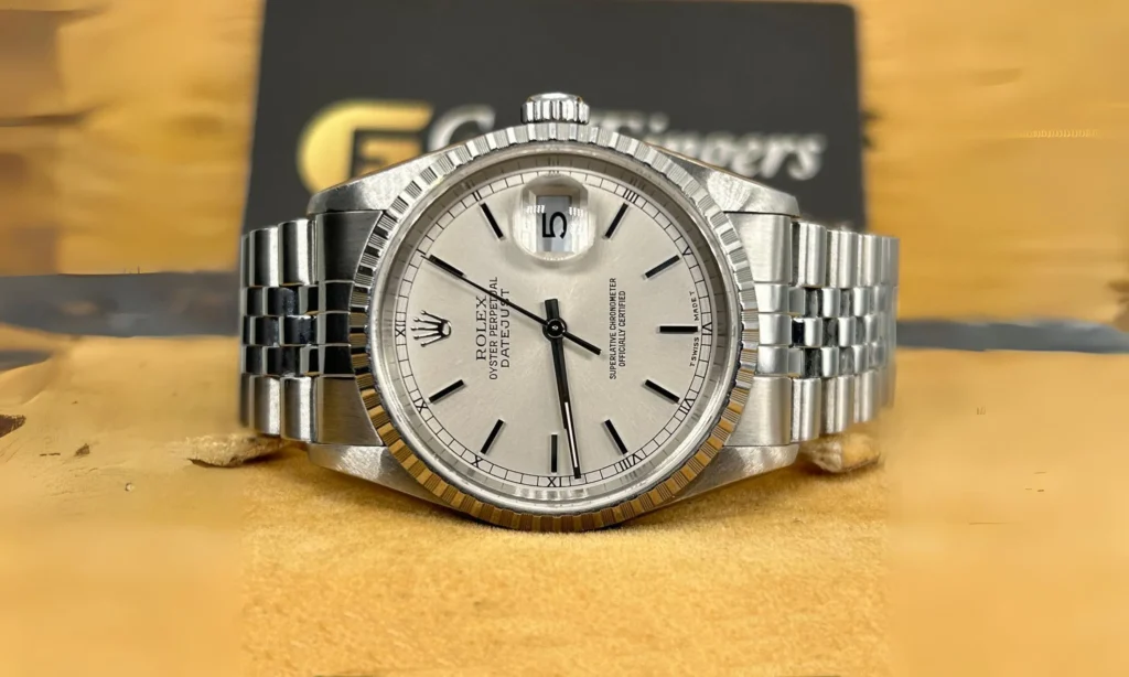 A silver Rolex Datejust 16220 watch with a white dial and date window, displayed on a tan surface.