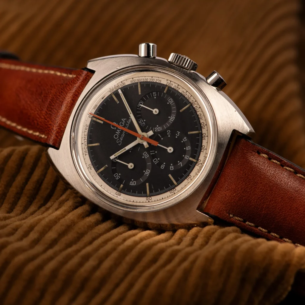 A vintage Omega Seamaster 145.006 chronograph watch with a brown leather strap rests elegantly on a corduroy fabric background.