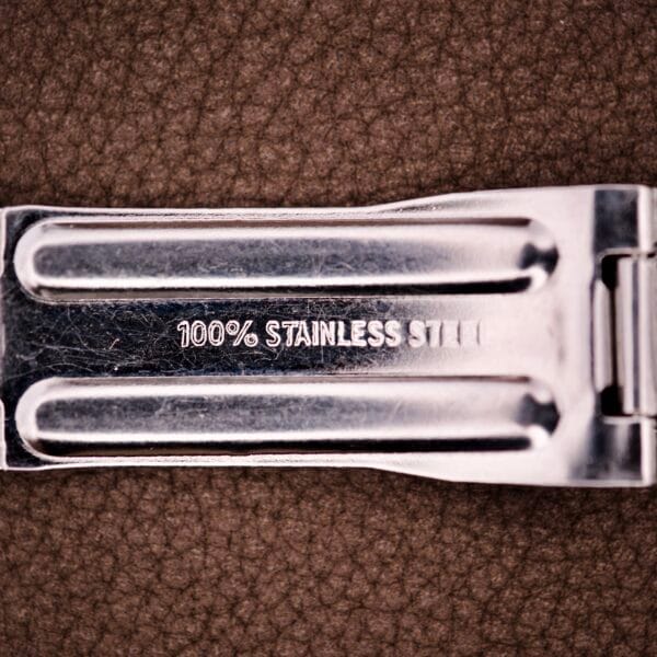 Close-up of an Enicar Sherpa Guide 600 "Pepsi" Automatic World Timer GMT Ref. 2342, featuring a metal part stamped 100% STAINLESS STEEL, set against a brown textured background.