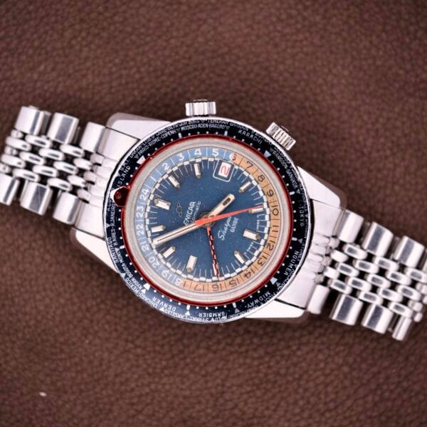 An Enicar Sherpa Guide 600 "Pepsi" Automatic World Timer GMT Ref. 2342 with a blue and red dial sits elegantly on a brown textured surface.