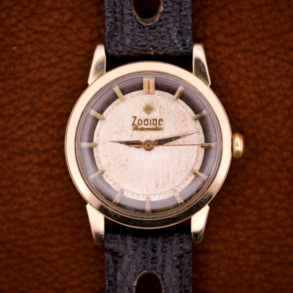 Zodiac Glorious Automatic Tuxedo Dial Ref. 688 Cal. 1361 wristwatch with gold case, cream dial, and black leather strap is beautifully presented against a brown background.