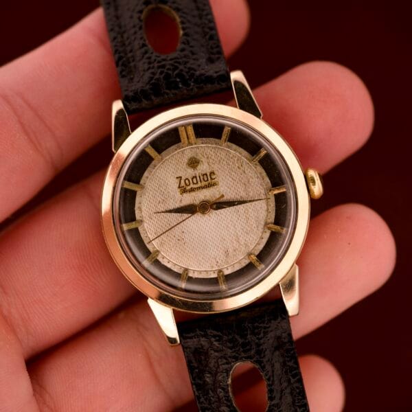 A hand holds a Zodiac Glorious Automatic Tuxedo Dial Ref. 688 wristwatch with a black leather strap, gold-tone case, and Cal. 1361 automatic movement.