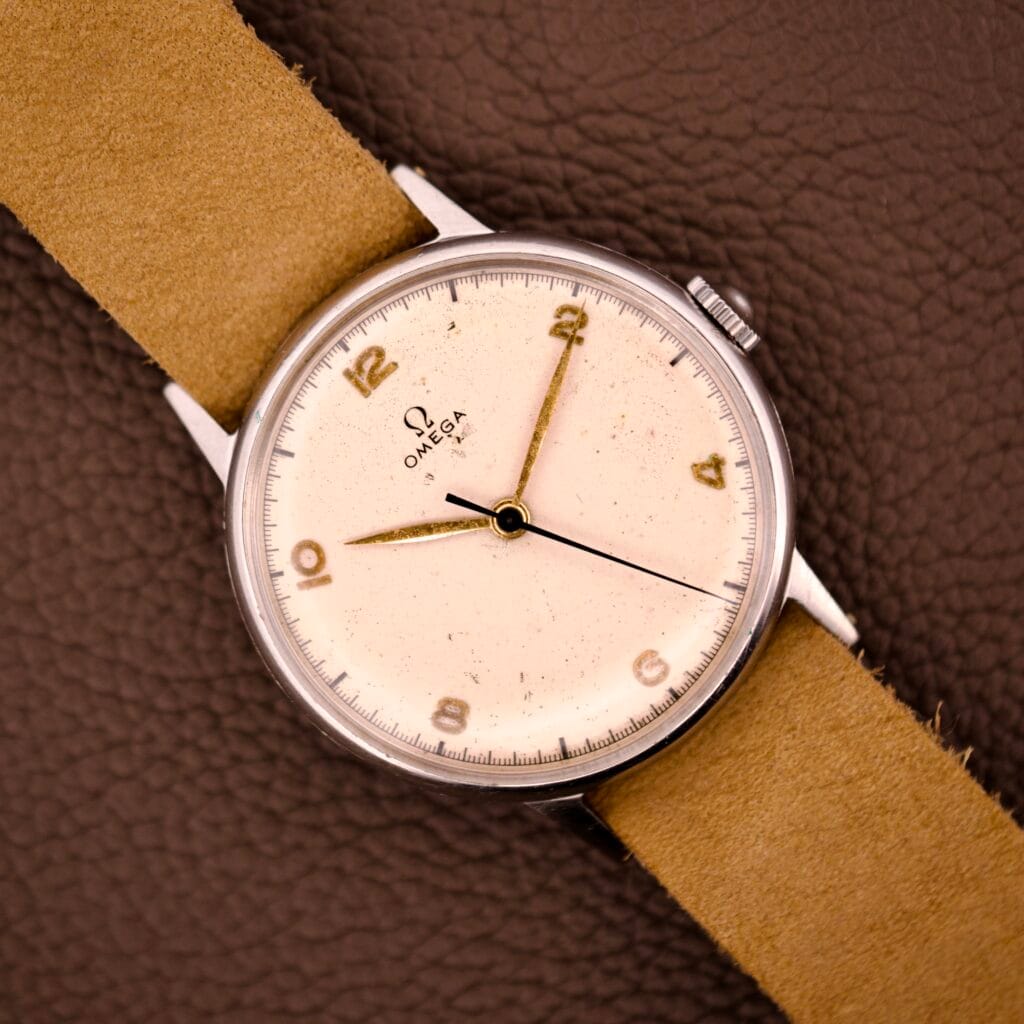 An Omega Ref. 2338 Jumbo wristwatch with a stainless steel case and beige strap rests on a brown textured surface.