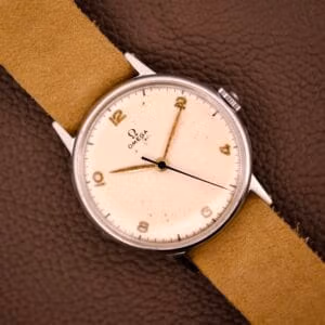 An Omega Ref. 2338 Jumbo wristwatch with a stainless steel case and beige strap rests on a brown textured surface.