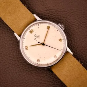 An Omega Ref. 2338 Jumbo wristwatch with a stainless steel case and beige strap rests on a brown textured surface.