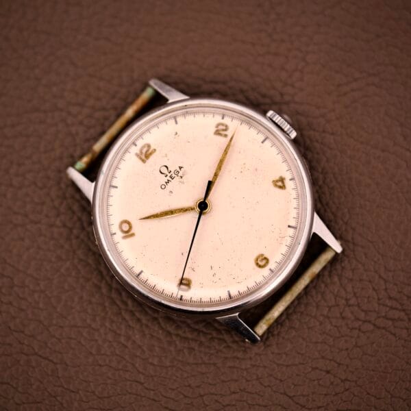 The Omega Ref. 2338 Jumbo features a cream dial with gold numerals, set in a classic stainless steel case with Caliber 30T2 SC, displayed on a brown textured surface.