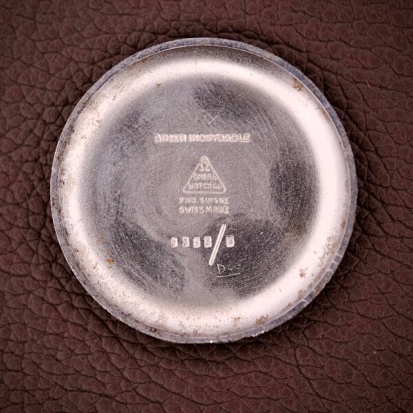 An Omega Ref. 2338 Jumbo Stainless Steel Case Back for Caliber 30T2 SC, with engraved Omega markings, is displayed on a textured brown surface.