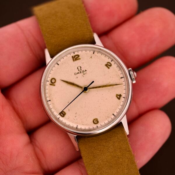 A hand holding an Omega Ref. 2338 Jumbo with a stainless steel case, beige dial, Caliber 30T2 SC movement, and an olive green suede strap.