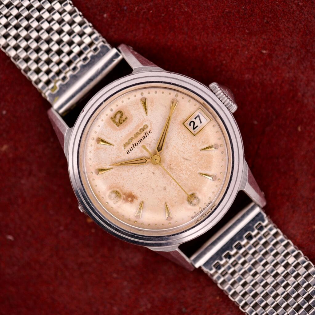 A vintage Movado Automatic Date Ref. 16291 wristwatch featuring a stainless steel mesh band and beige dial, with date 27 shown on a red background; powered by Bumper Cal. 224A movement.