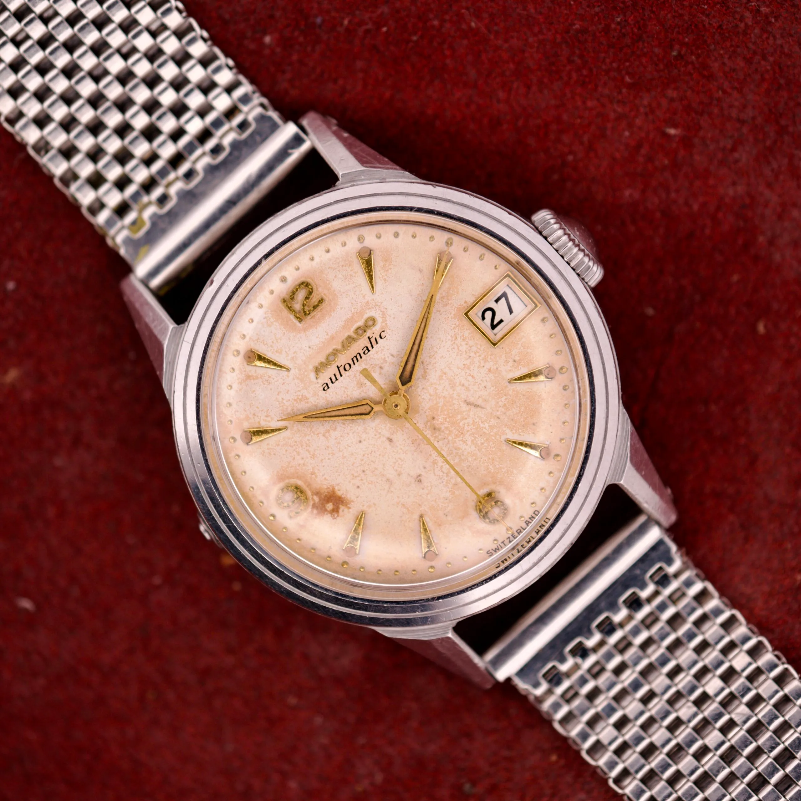 A vintage Movado Automatic Date Ref. 16291 wristwatch featuring a stainless steel mesh band and beige dial, with date 27 shown on a red background; powered by Bumper Cal. 224A movement.