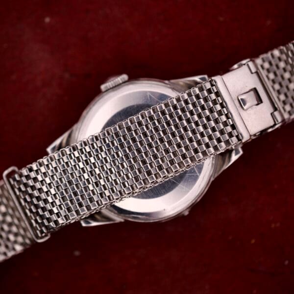 A silver stainless steel mesh band partially covers the back of a round Movado Automatic Date Ref. 16291 Bumper Cal. 224A watch, shown on a red background.