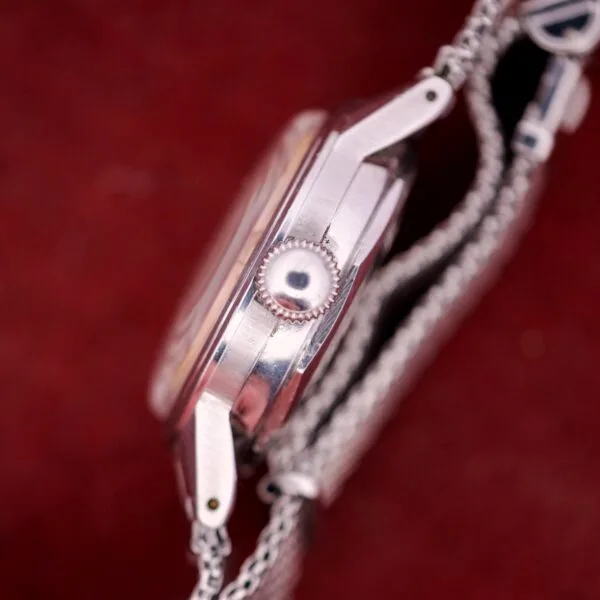 Side view of the Movado Automatic Date Ref. 16291, featuring a stainless steel case and metal mesh band, set against a red background.