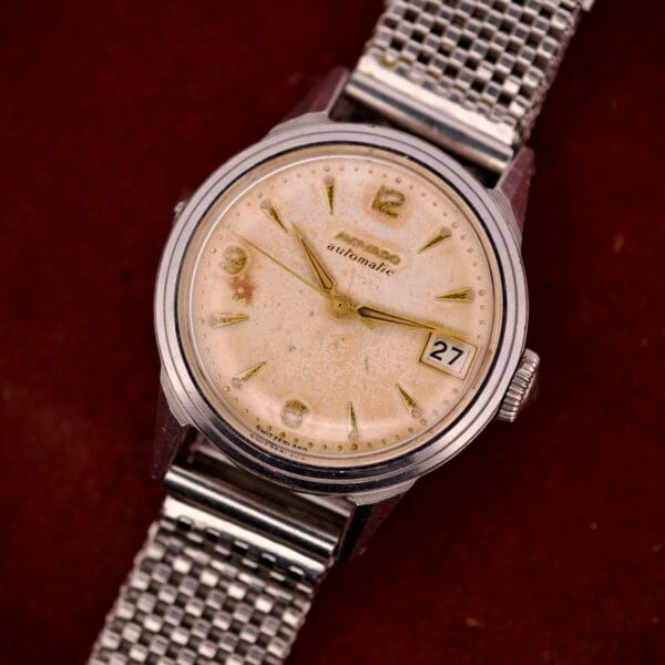 A vintage Movado Automatic Date Ref. 16291 with a stainless steel band, yellowed dial, and date window is displayed against a red backdrop.