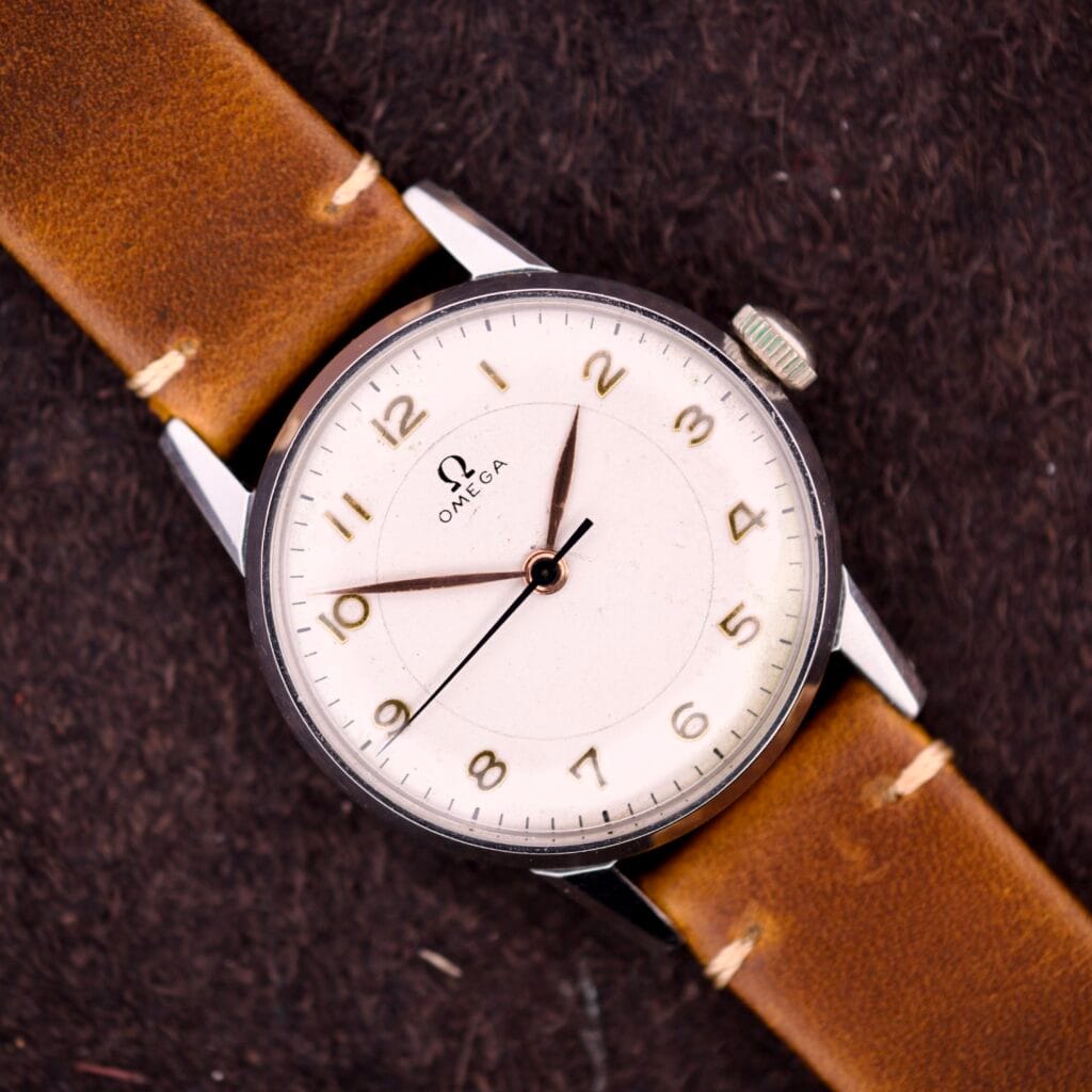 An Omega Ref. 2496 Arabic Numerals Stainless Steel Case Cal. 30T2 SC wristwatch with a brown leather strap rests on a dark textured surface.