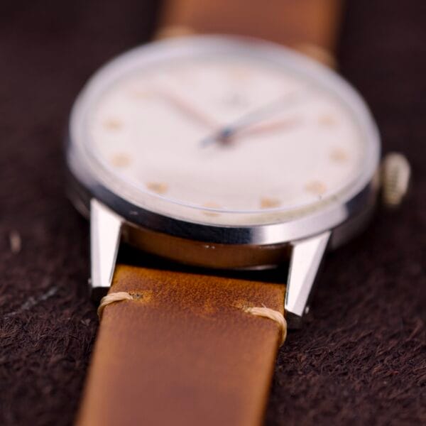 A close-up of an Omega Ref. 2496 Arabic Numerals with a stainless steel case, Cal. 30T2 SC movement, worn brown leather strap, and blurred white dial featuring Arabic numerals.
