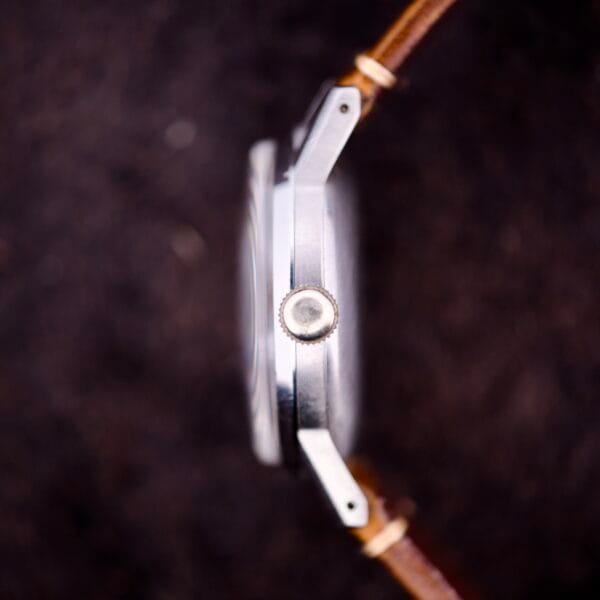 Side view of an Omega Ref. 2496 Arabic Numerals Stainless Steel Case Cal. 30T2 SC wristwatch with a brown leather strap, highlighting the crown against a dark background.