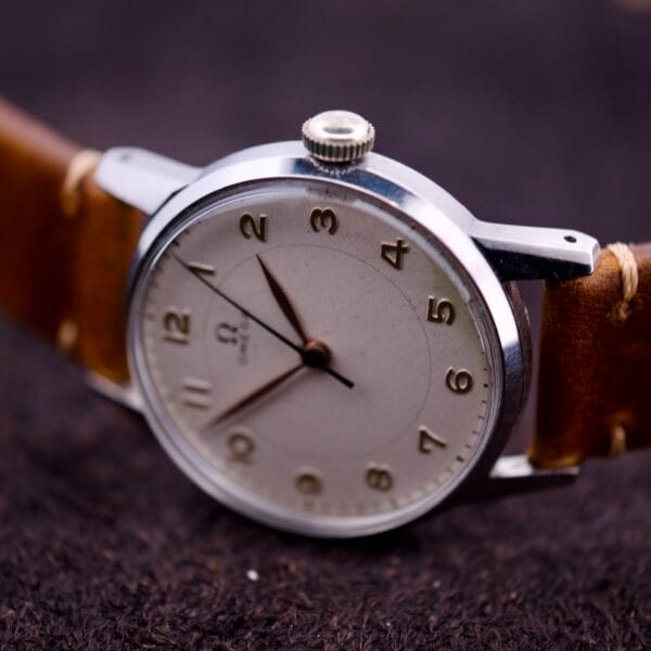 An Omega Ref. 2496 Arabic Numerals watch with a stainless steel case, Cal. 30T2 SC movement, and brown leather strap rests on a dark textured surface.